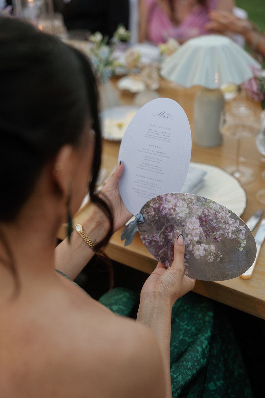 Wedding guest holds oval menu card with floral design at elegantly set outdoor reception table.