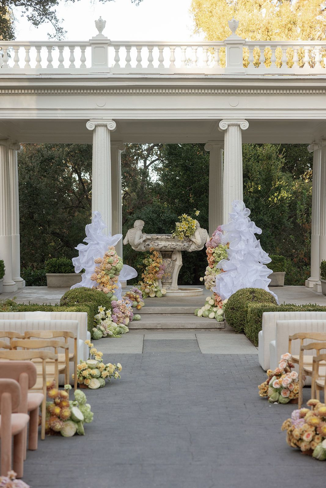 Elegant outdoor wedding ceremony with floral arrangements, white columns, and a stone statue altar in a garden setting.