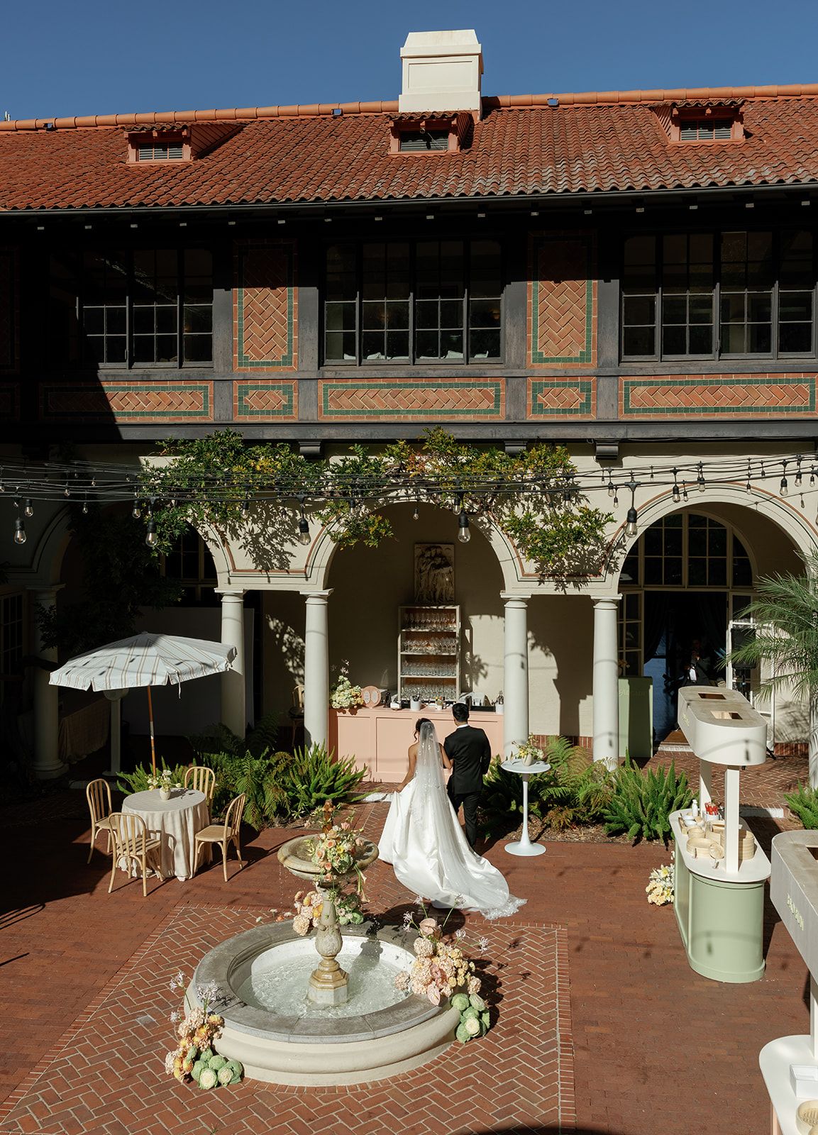 Bride in white gown stands by fountain in elegant Spanish-style courtyard with arched colonnade during outdoor wedding reception.