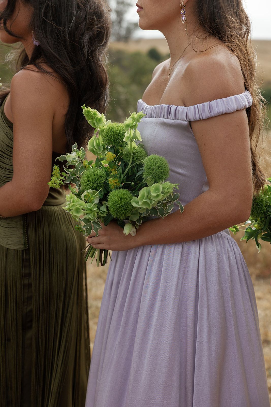Two bridesmaids in lavender and olive dresses holding green bouquets at an outdoor wedding ceremony.