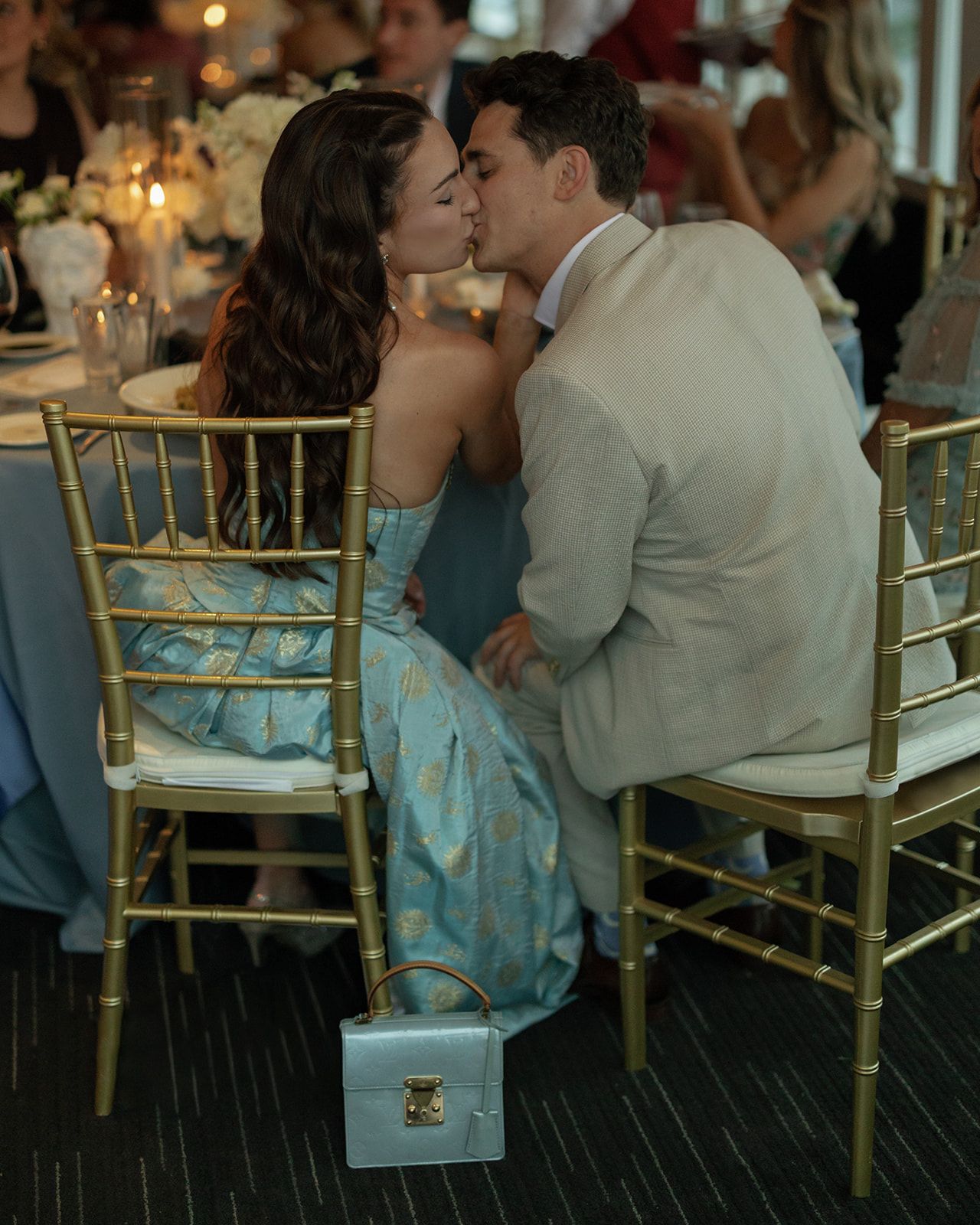 A couple shares an intimate moment while sitting at an elegant wedding reception with gold chairs and decorative lighting.