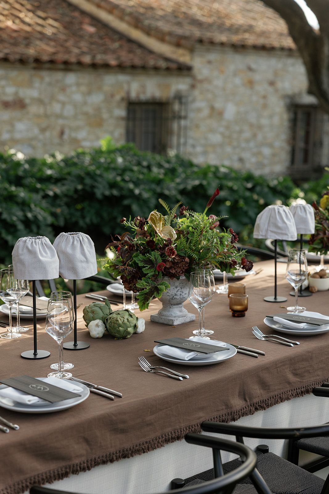 Elegant outdoor table setting with brown linen tablecloth, white plates, and natural greenery centerpiece at rustic venue.