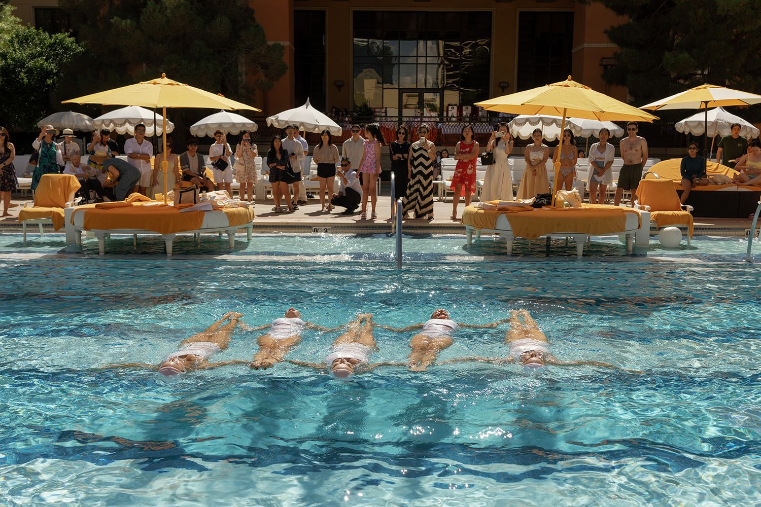 Synchronized swimmers perform in a luxury hotel pool surrounded by yellow umbrellas and lounging spectators on a sunny day.