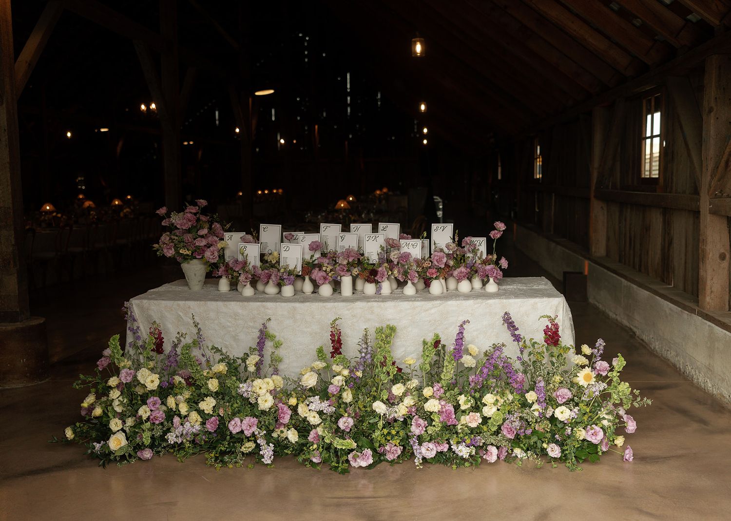 Elegant barn wedding reception table adorned with lush pink and purple floral arrangements and white candles in rustic setting.