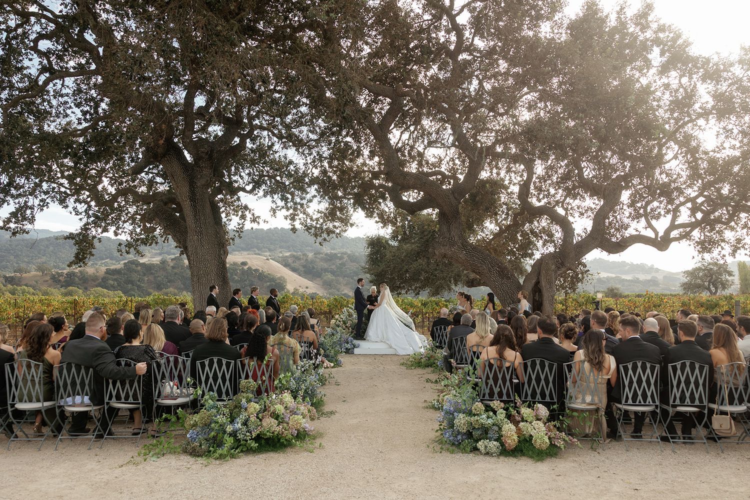 Outdoor wedding ceremony under oak trees with guests seated on black chairs amid rolling hills and floral arrangements.