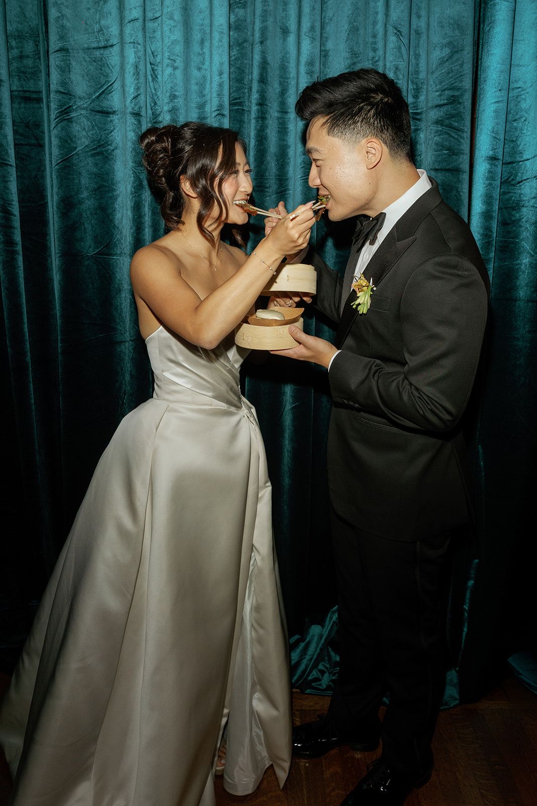 Bride and groom sharing cake at wedding reception, wearing elegant white gown and black tuxedo, teal velvet curtain backdrop.