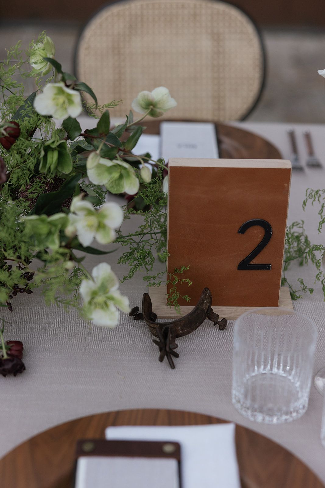 Rustic wedding table setting with wooden number 2 sign, hellebore flowers, and elegant glassware on white tablecloth.
