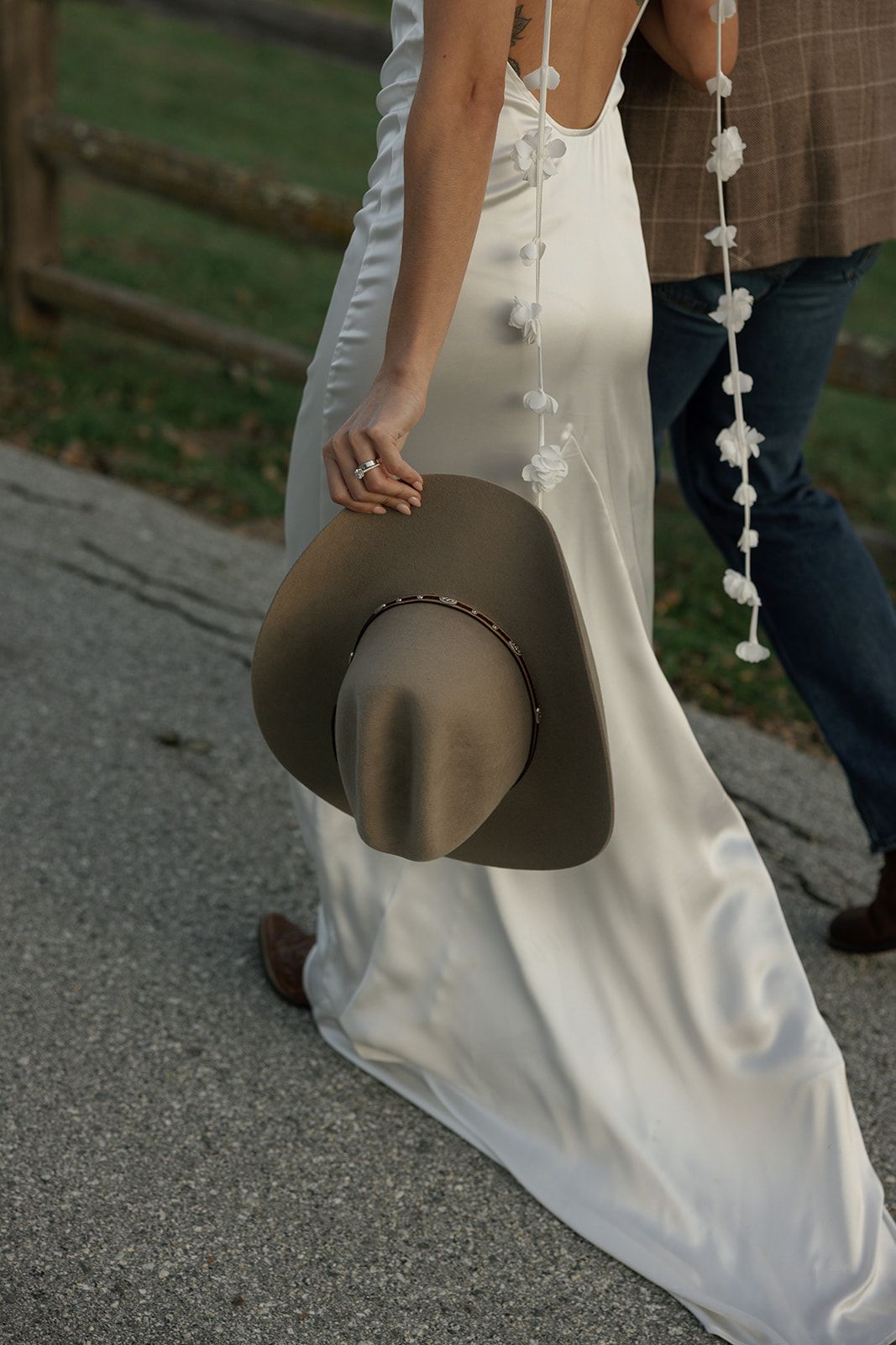 A person in a flowing white dress holds a brown wide-brimmed hat while walking on a paved path.