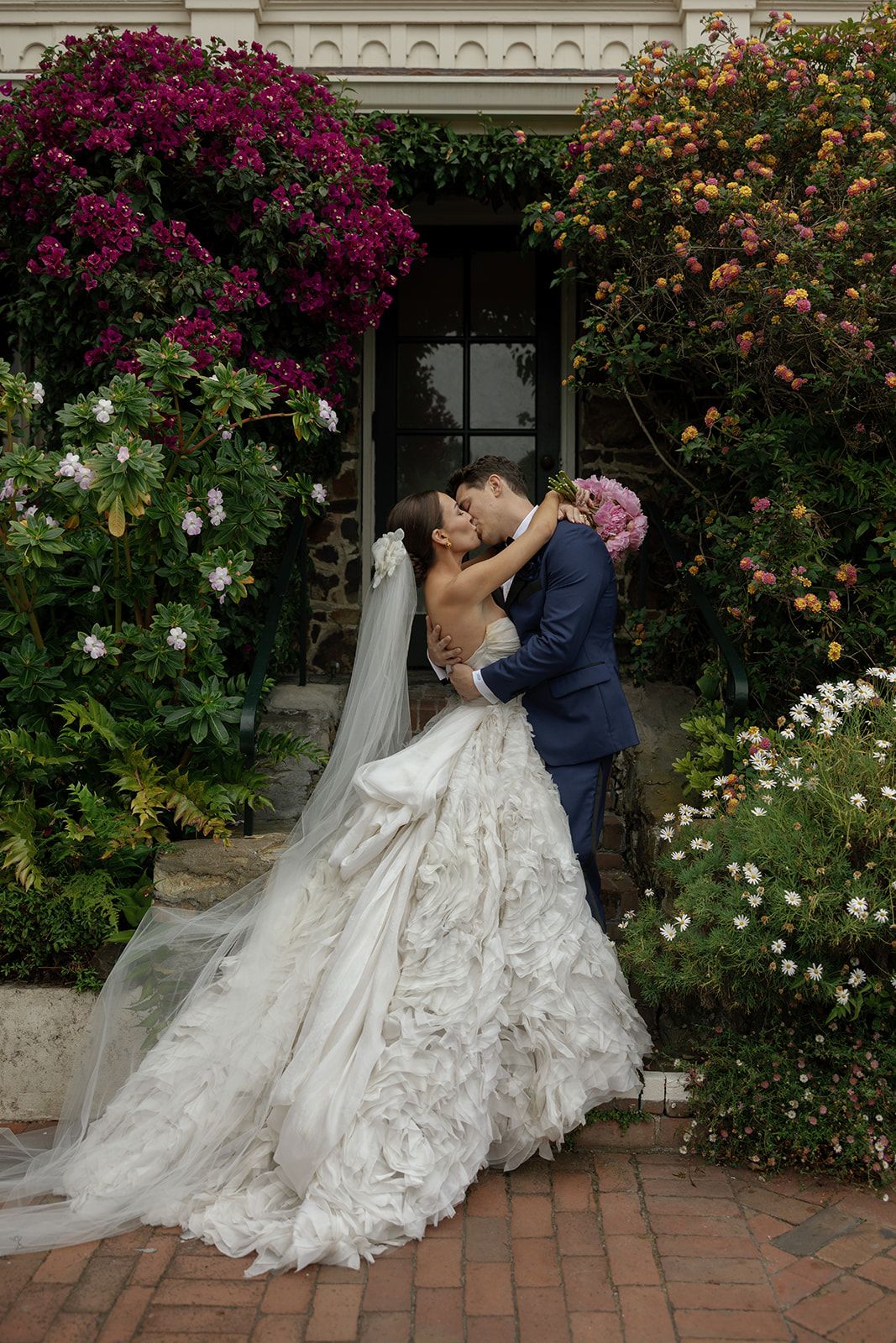 A romantic wedding couple embrace in front of a vibrant garden backdrop with magenta bougainvillea and white roses.