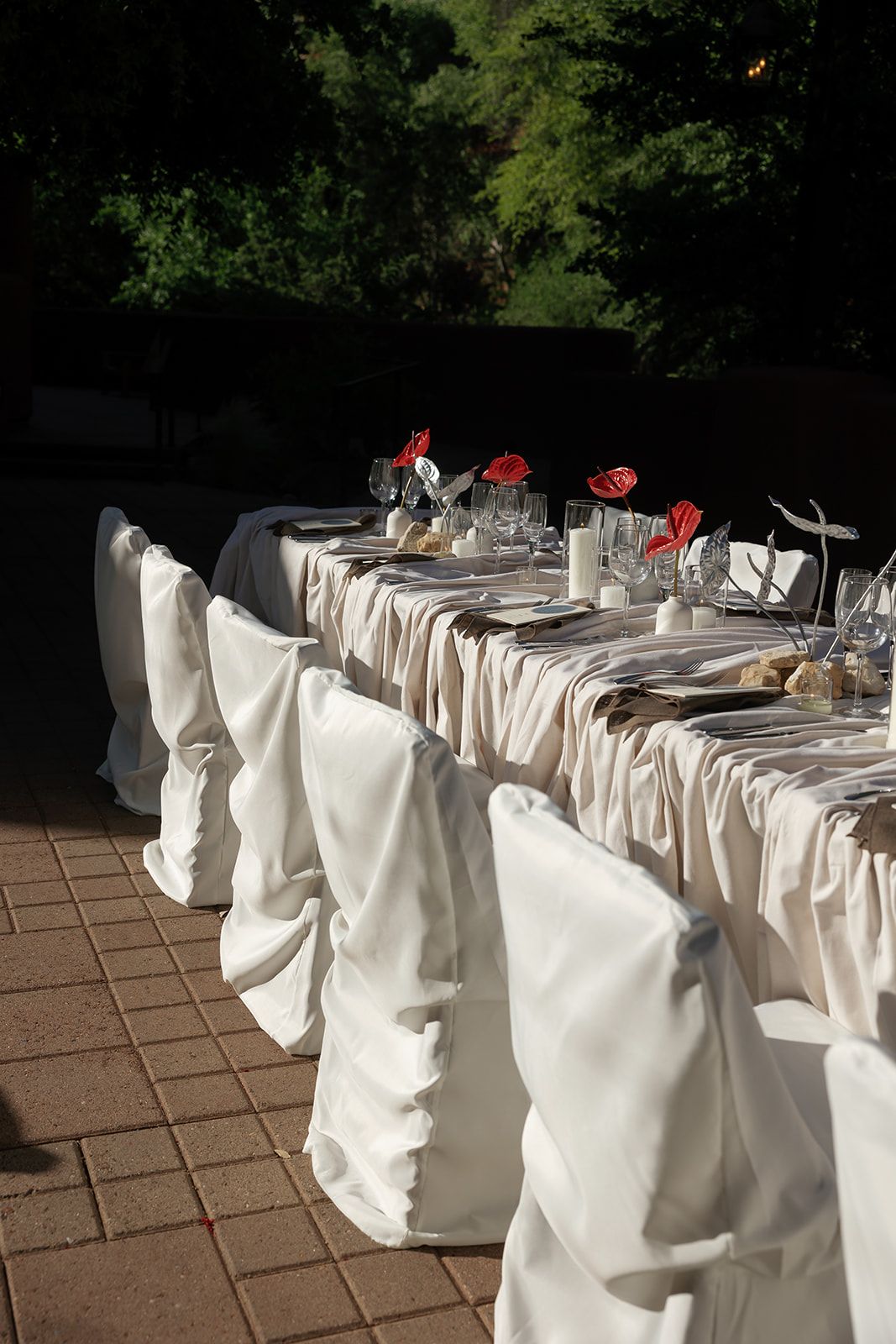 An elegant wedding table setup with white chair covers, silver tableware, and red floral centerpieces arranged outdoors.