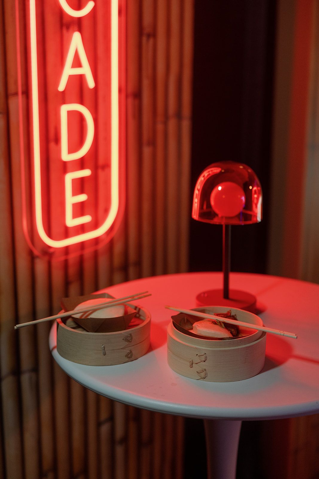 Two bamboo dim sum steamers with chopsticks on a white table under red neon arcade sign with glowing lamp.