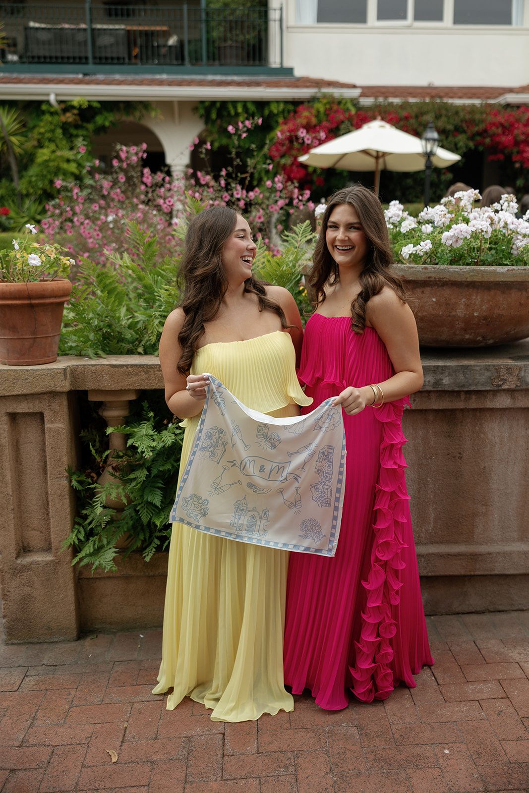 Two women in yellow and pink maxi dresses share a laugh while holding lace fabric in a garden setting with blooming flowers.