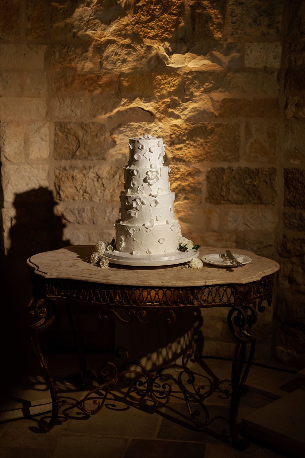 A white tiered wedding cake displayed on a wrought iron stand in a rustic stone cellar with dramatic lighting.