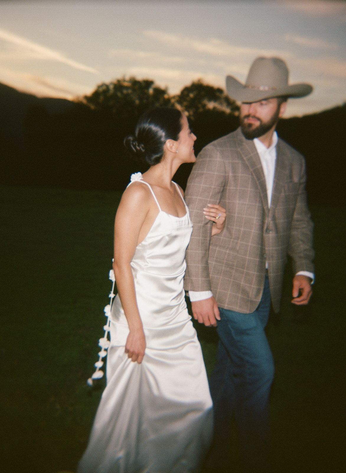 A couple in western attire dances together at sunset during a rustic outdoor wedding celebration.