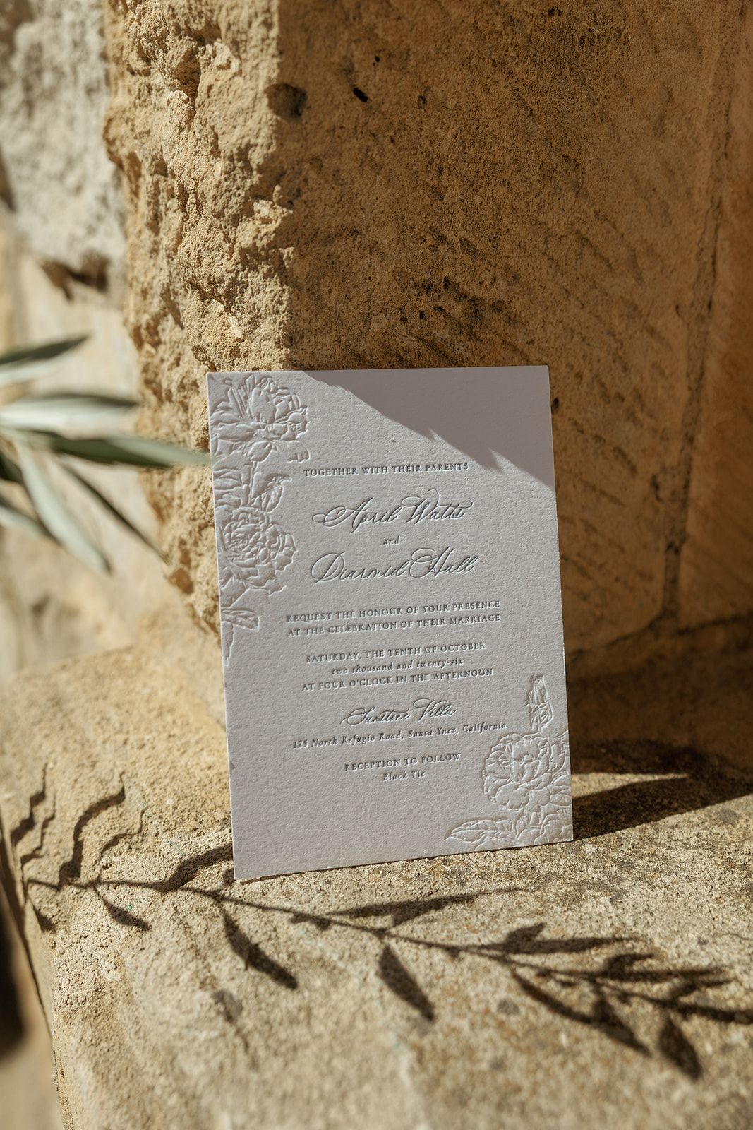 Elegant letterpress wedding invitation with floral embossing leaning against rustic stone wall in natural sunlight.