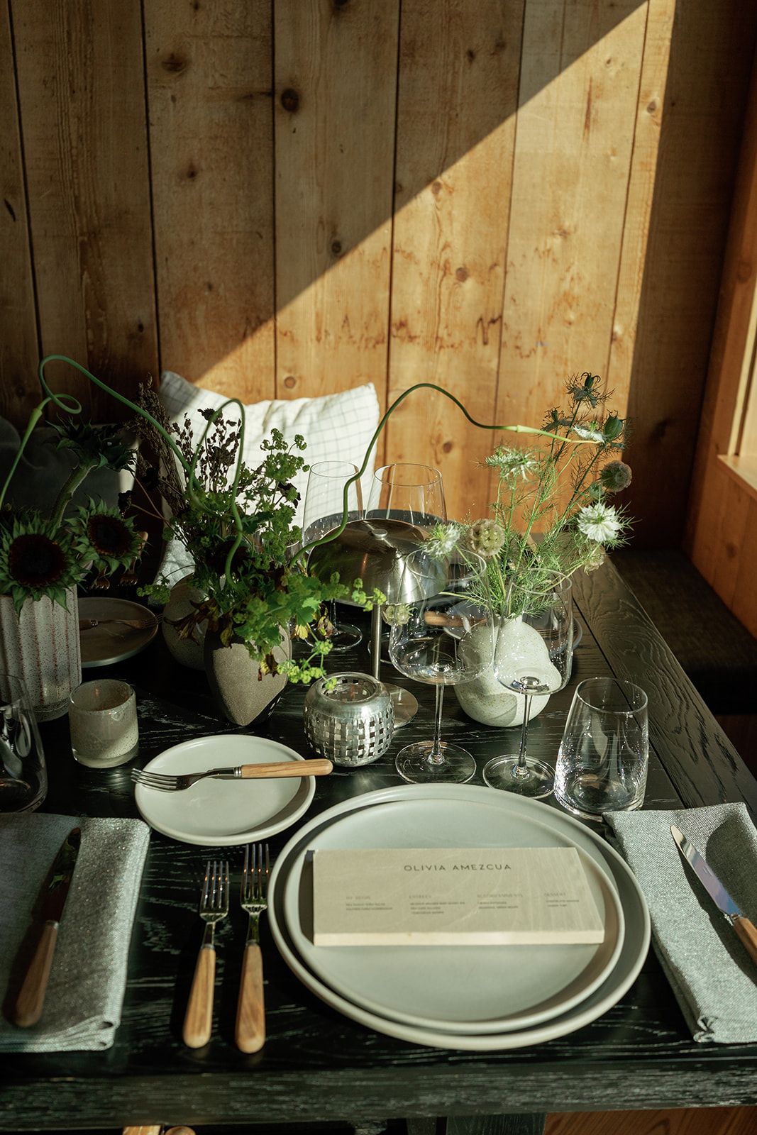 Rustic wooden table set with ceramic plates, menu card, wildflowers, and glassware in warm natural light.