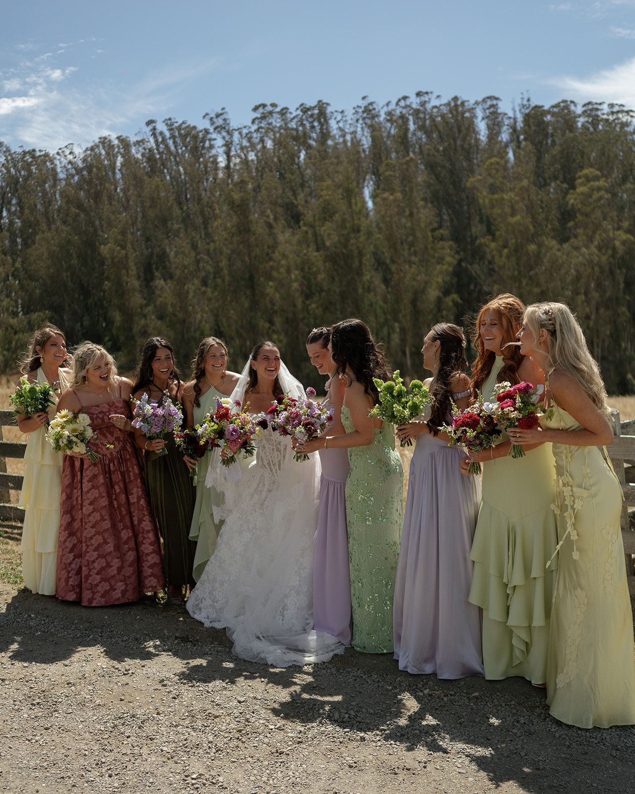 Bride in white gown stands with bridesmaids in mismatched colorful dresses holding floral bouquets outdoors.