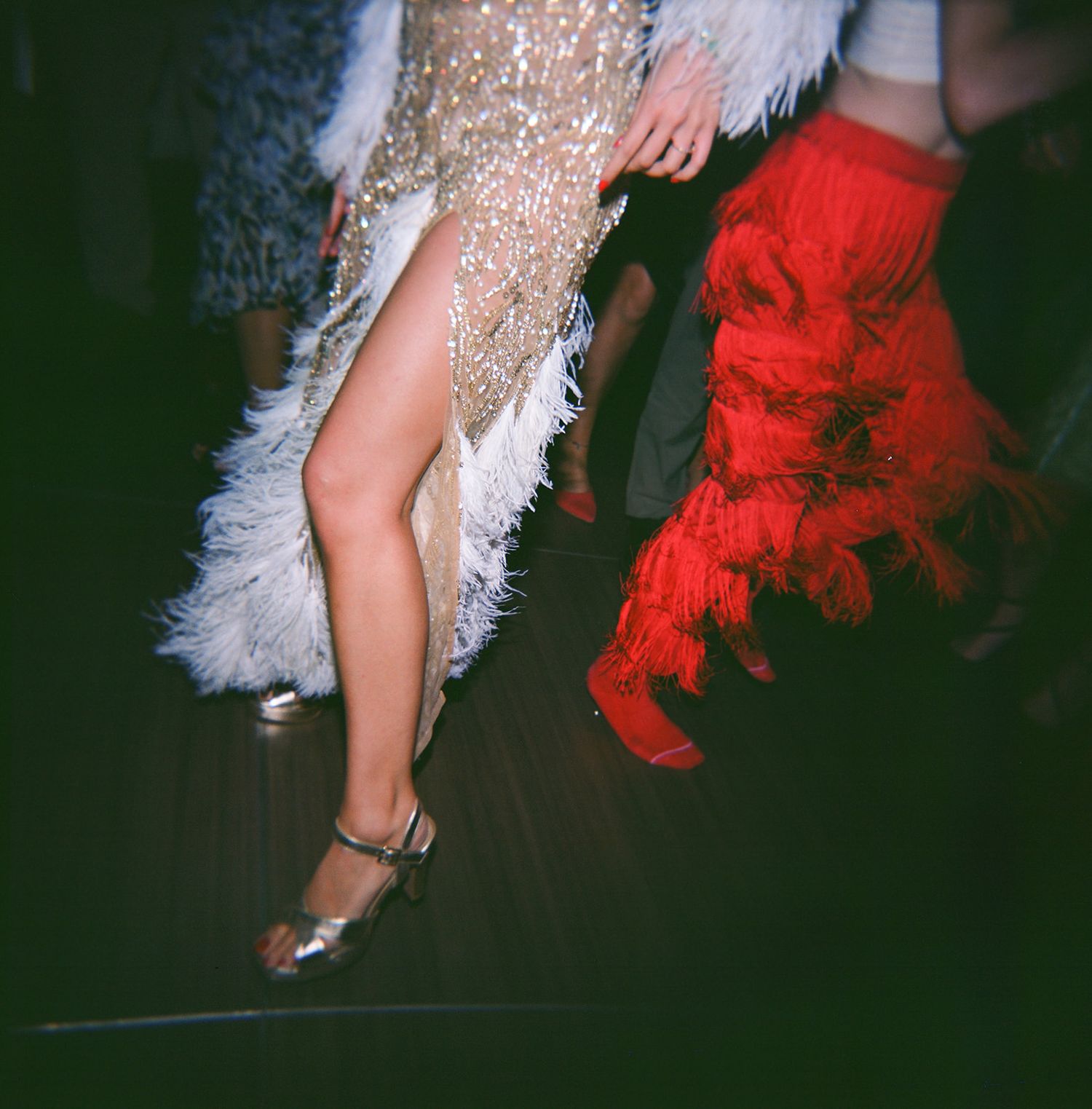 Dancing legs in sparkly silver and red dresses with feather details move across a dark dance floor.