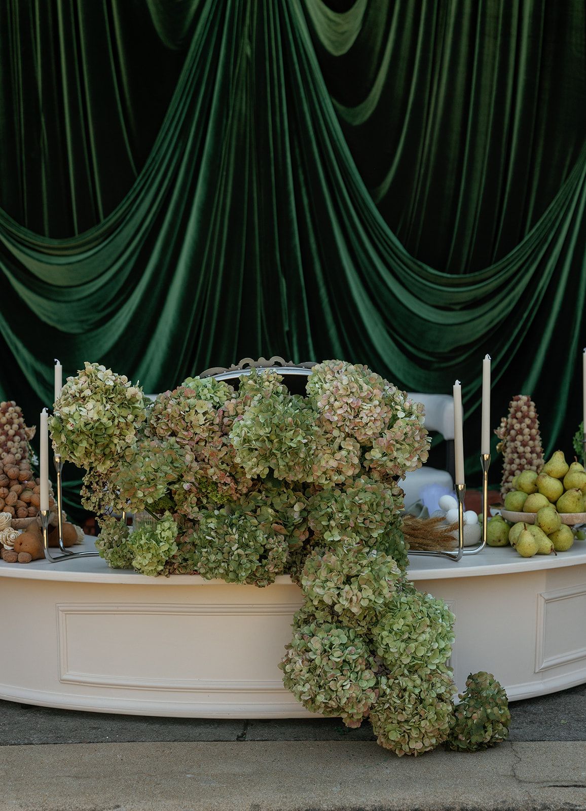 Dried hydrangea flowers arranged in a curved white ceramic vessel against a dark green draped backdrop.