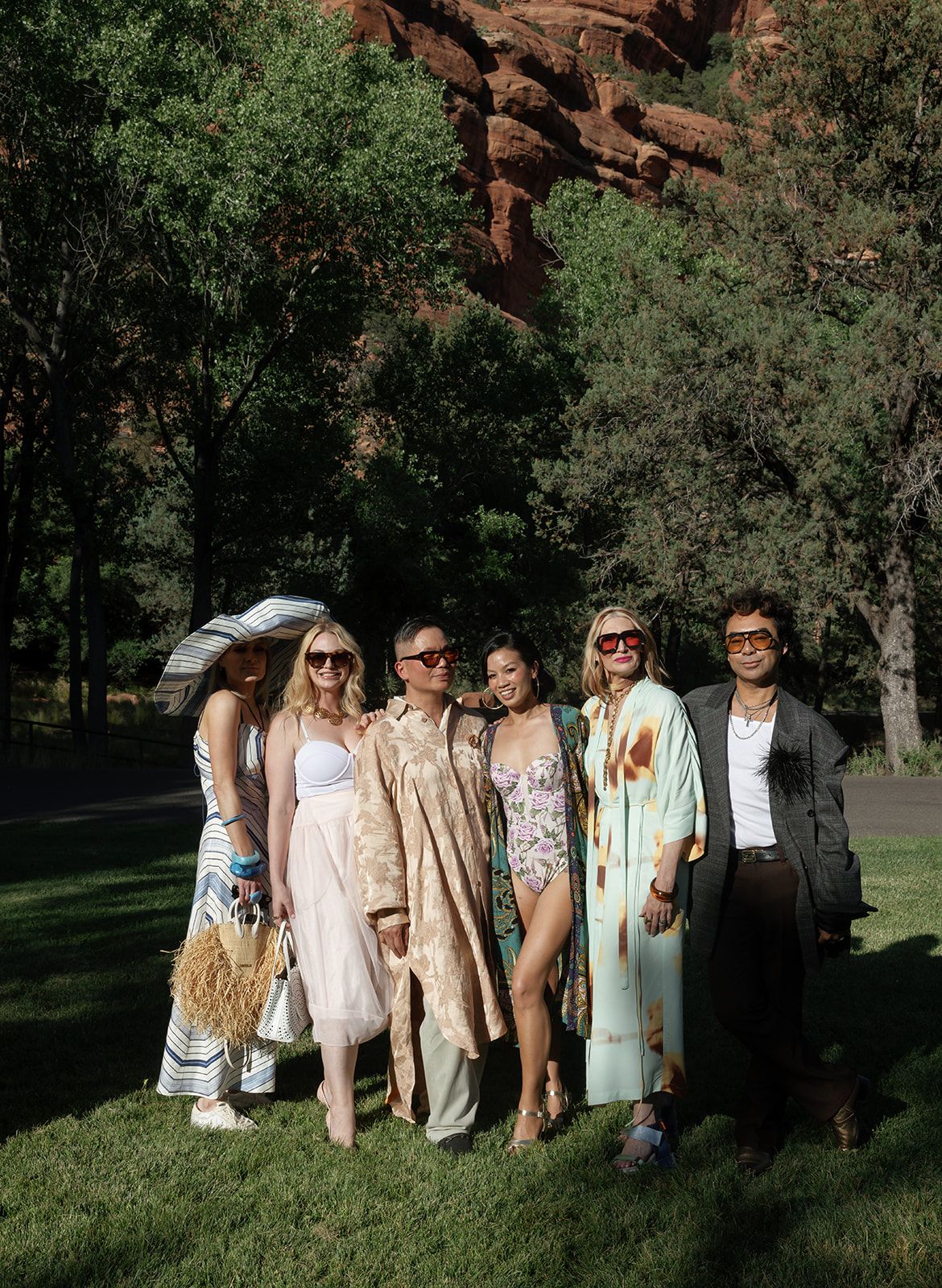 A stylishly dressed group poses together on a grassy lawn with red rock formations and trees in the background.