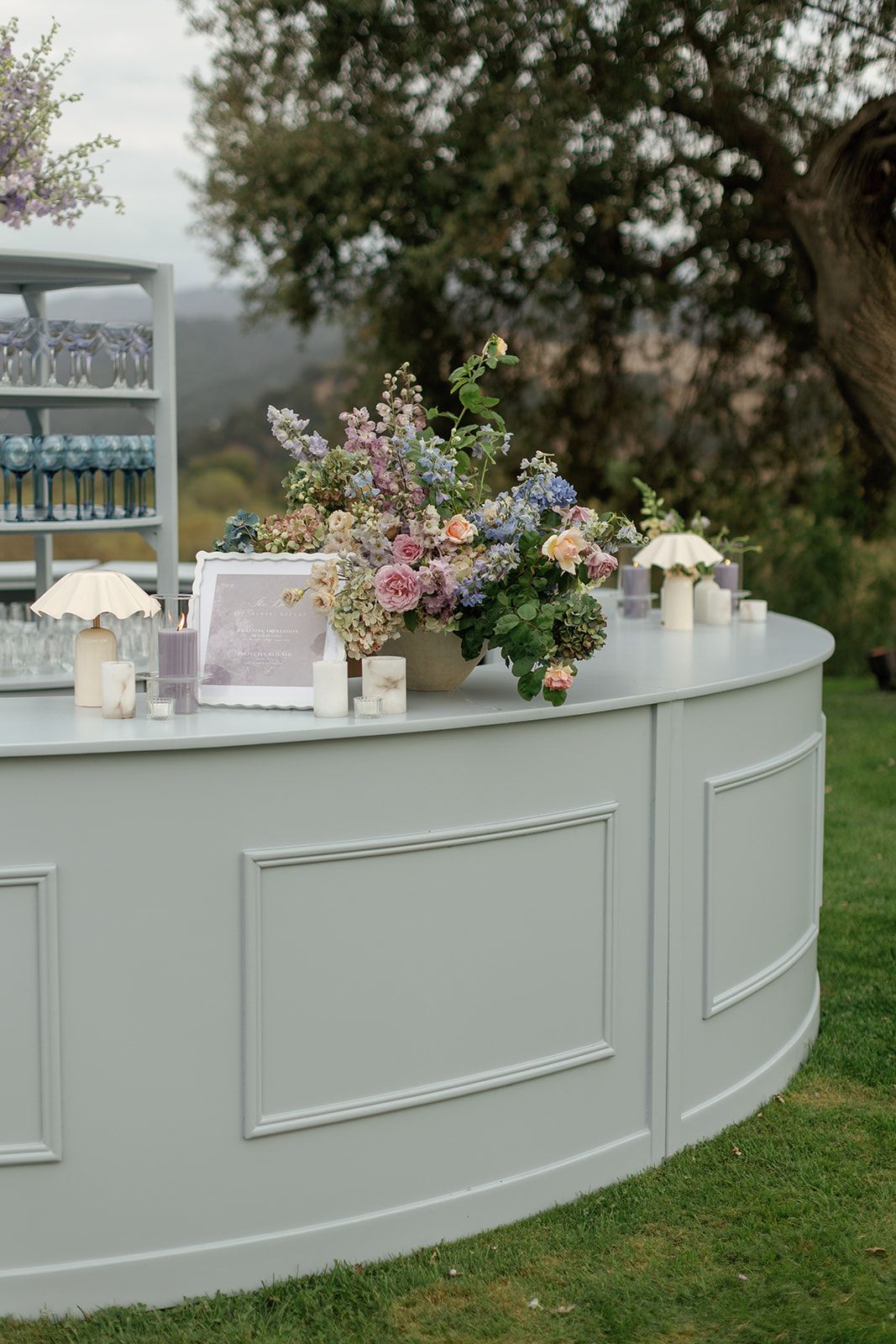 Elegant outdoor wedding bar with sage green paneling, floral arrangements in pink and blue, candles, and glassware display.