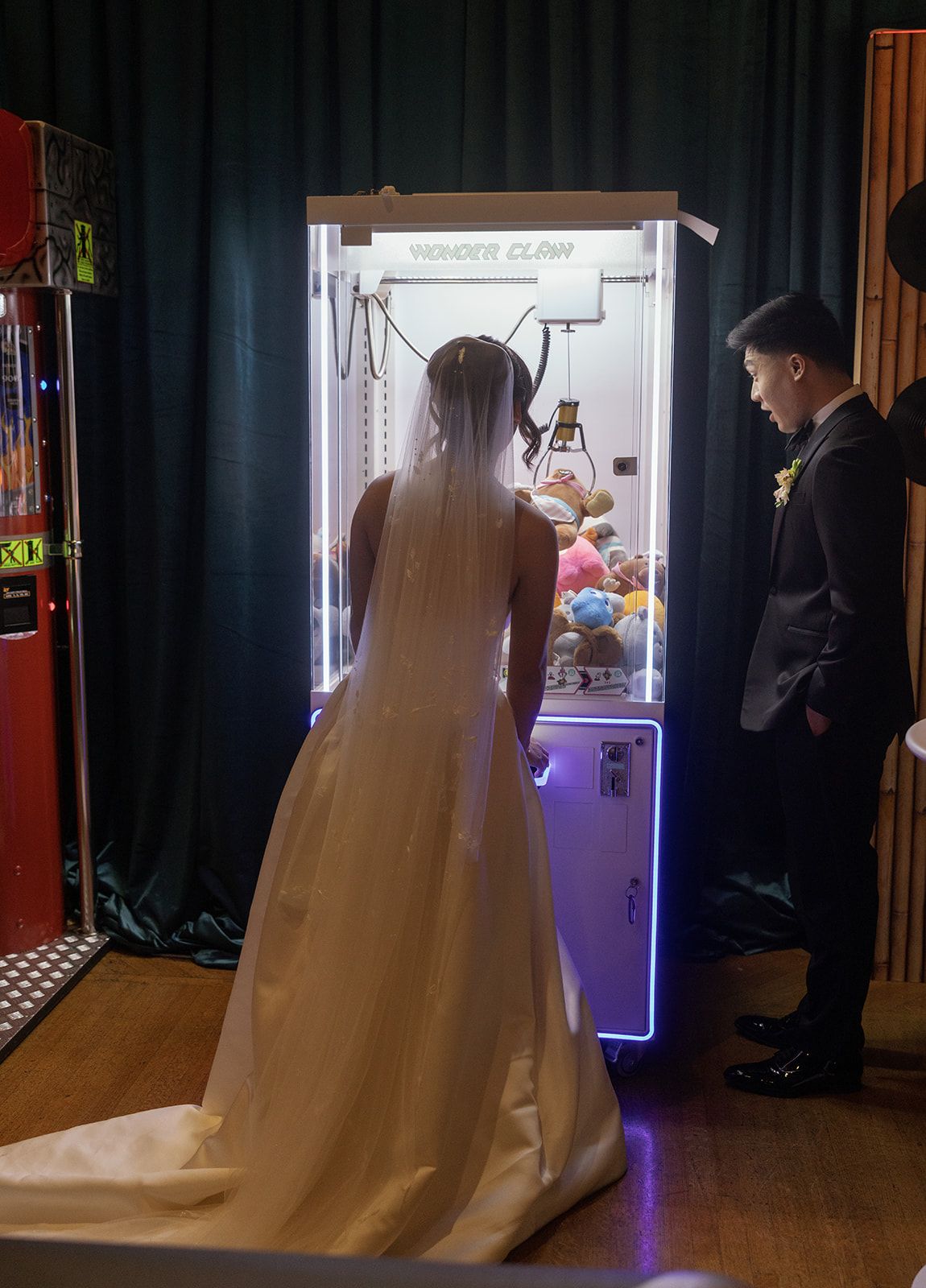 Bride in white wedding gown and groom in suit playing a claw machine arcade game at their wedding reception.