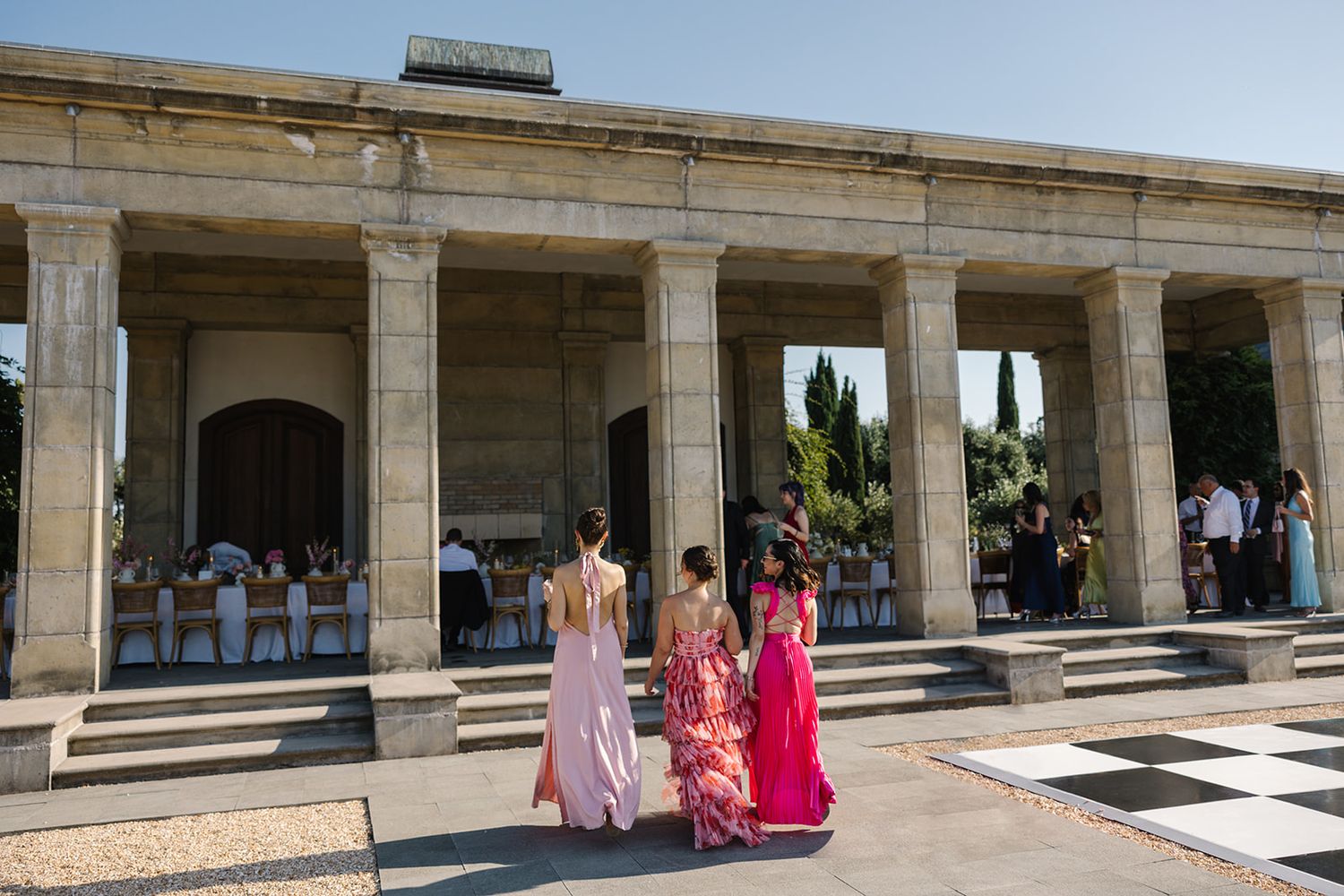 Wedding guests in colorful dresses walking through elegant stone colonnade at outdoor venue.