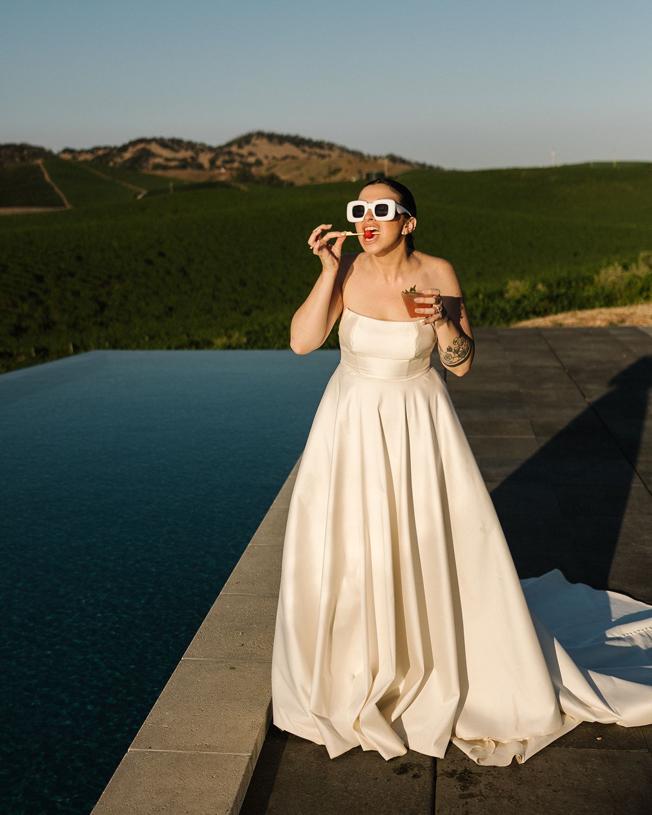 Bride in white gown and retro sunglasses enjoying refreshments by infinity pool.