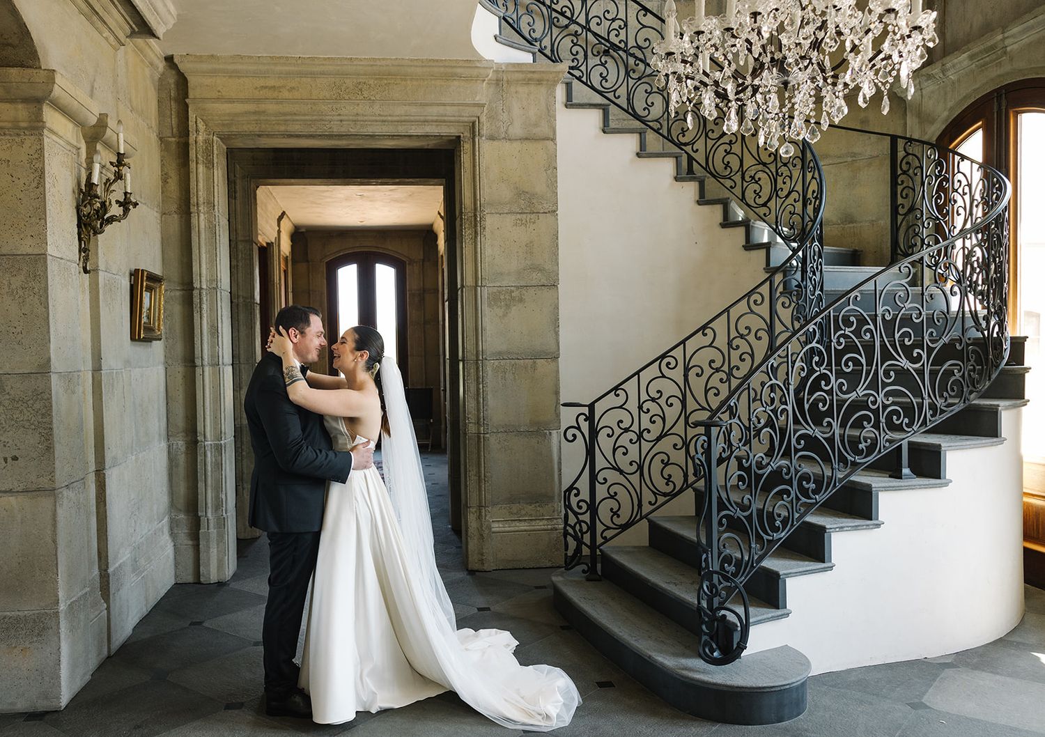 A romantic wedding moment captured in an elegant stone hallway with ornate wrought iron staircase and chandelier.
