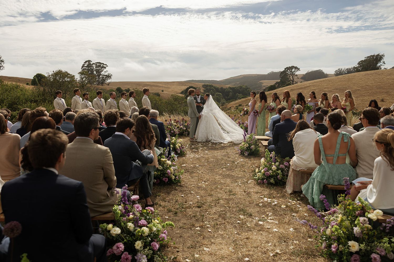 Outdoor wedding ceremony in golden hills with floral aisle, bride and groom at altar, guests seated, bridesmaids in green.