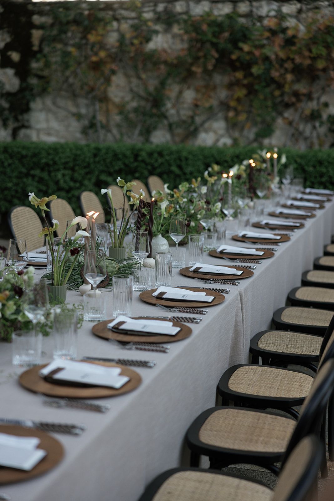 Elegant outdoor dining table setting with white linens, woven chairs, and minimalist place settings against a natural backdrop.