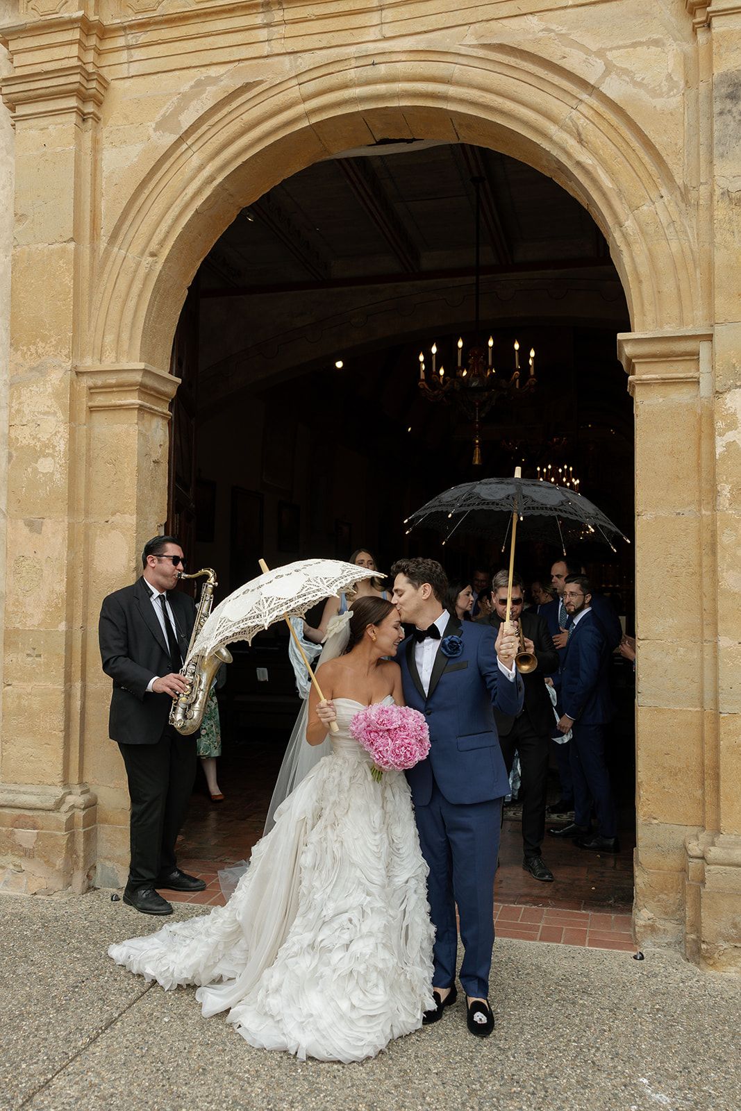 A couple exits a grand stone church archway holding white lace parasols at their outdoor wedding ceremony.