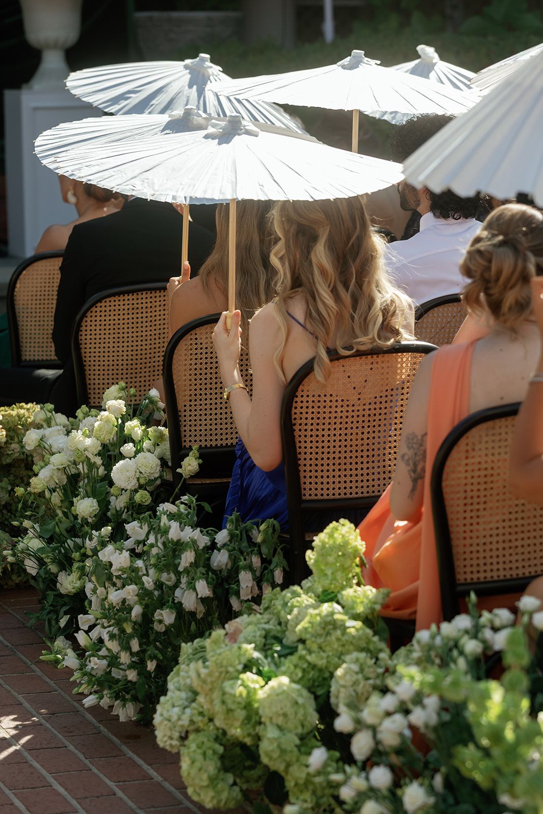Guests sit in cane chairs holding white parasols at an outdoor event with hydrangea and floral arrangements.