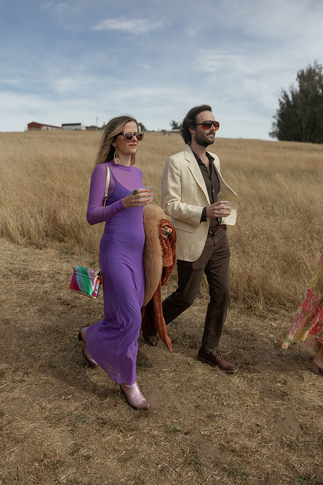 A woman in a purple maxi dress and a man in a cream blazer walk through a golden field wearing red sunglasses.