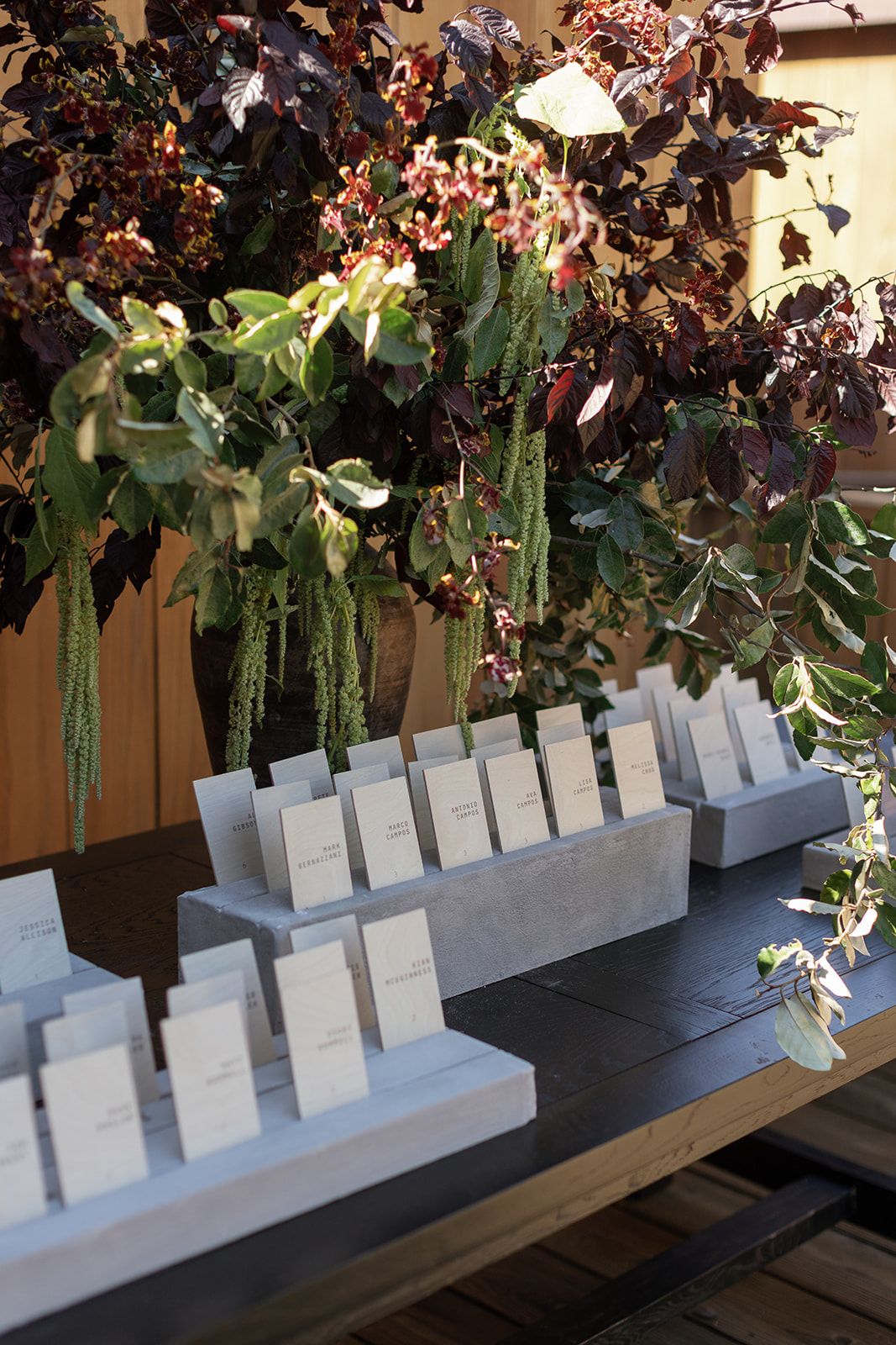 Elegant escort card display on dark table with tiered marble holders, surrounded by lush floral arrangements with burgundy foliage.