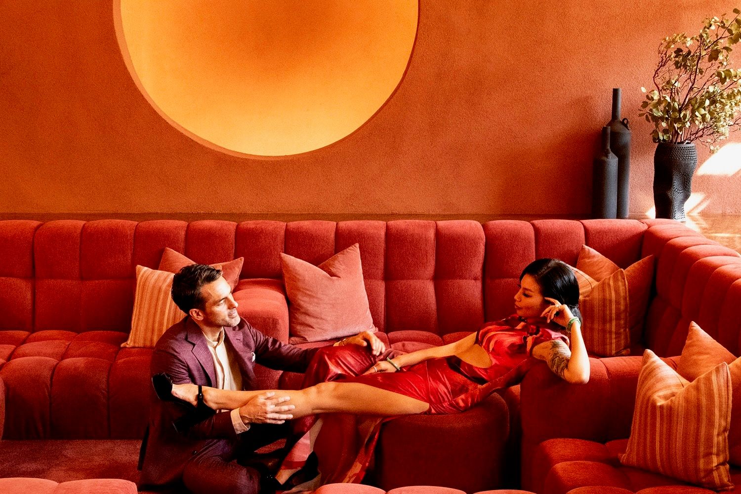 Two people sit in conversation on a red velvet couch beneath a large orange circular light fixture in a moody lounge setting.