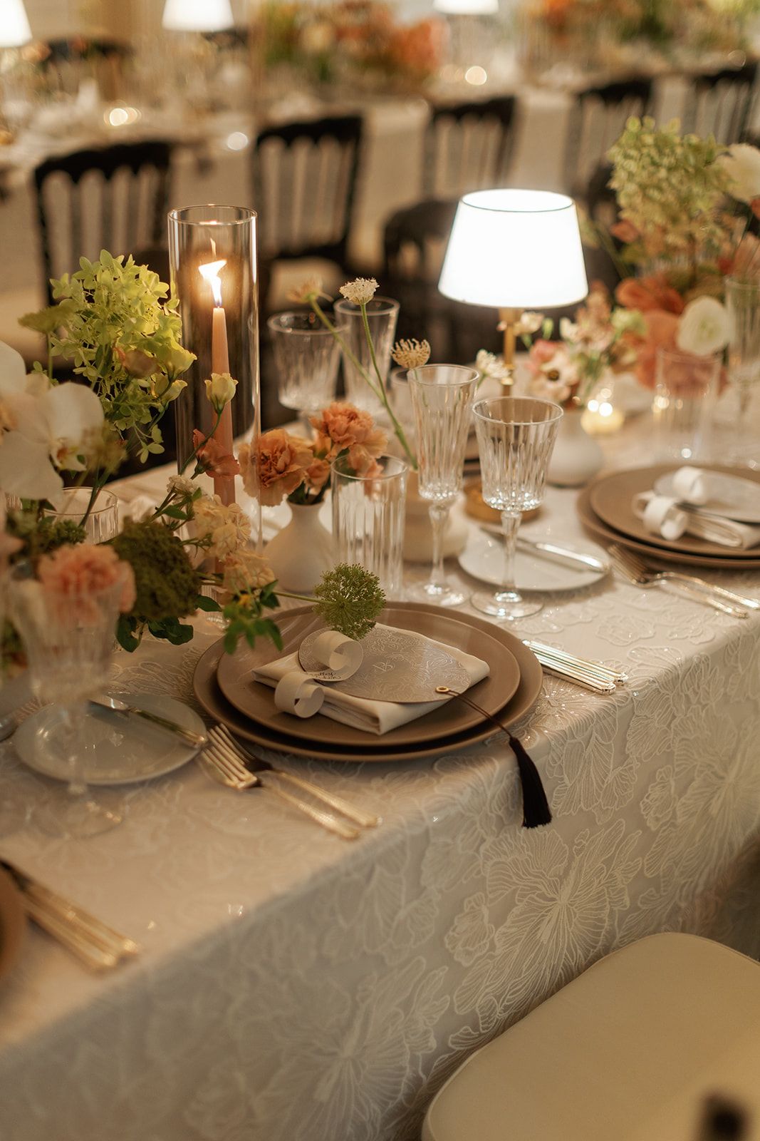 Elegant wedding reception table set with crystal glasses, candles, peach floral arrangements, and white embroidered linen tablecloth.