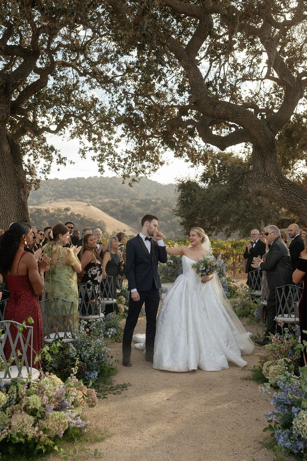 Bride and groom walk down outdoor aisle under oak tree at vineyard wedding with hydrangea floral arrangements.