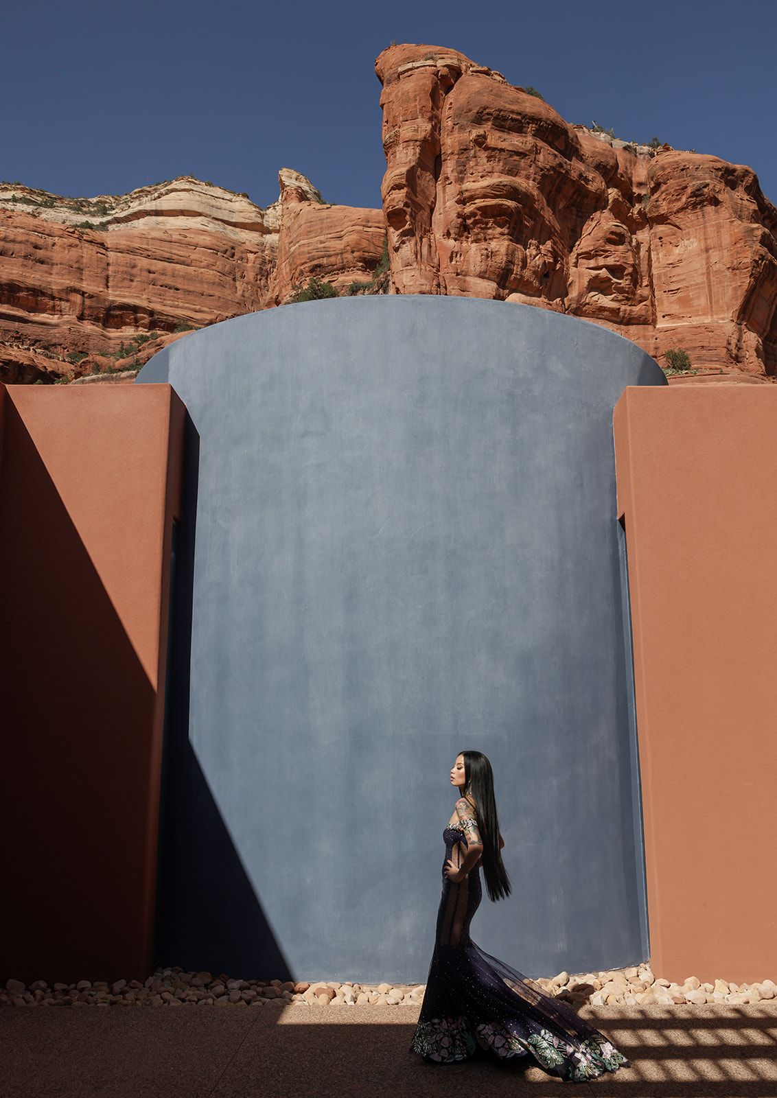 A figure stands against a curved blue wall in a desert landscape with red rock formations and sand dunes.