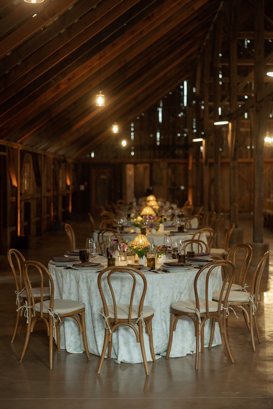 Rustic barn wedding reception with bentwood chairs, white linen tables, floral centerpieces, and warm Edison bulb string lights.