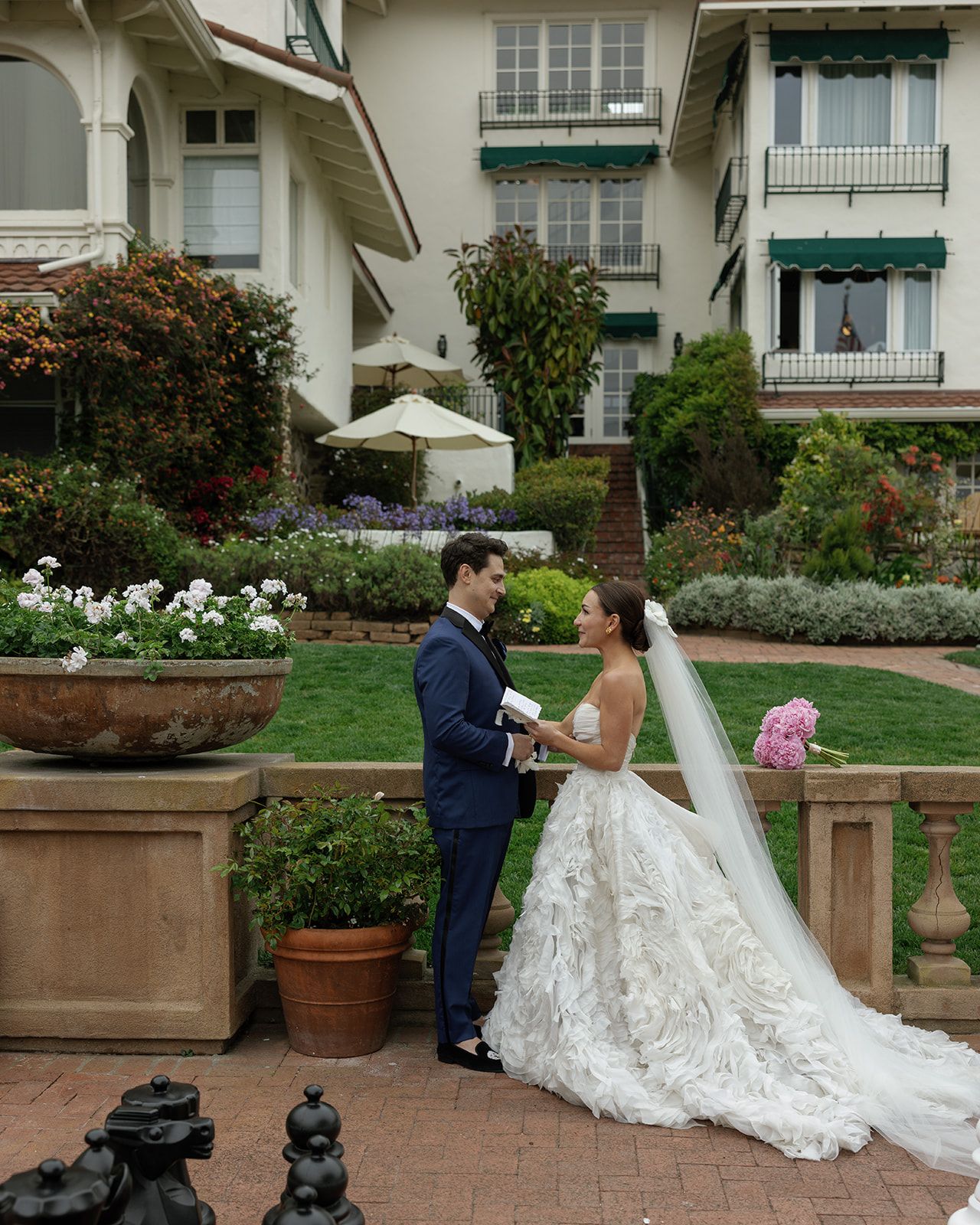 A bride and groom share a romantic first look moment in a garden setting with potted plants and white architecture.