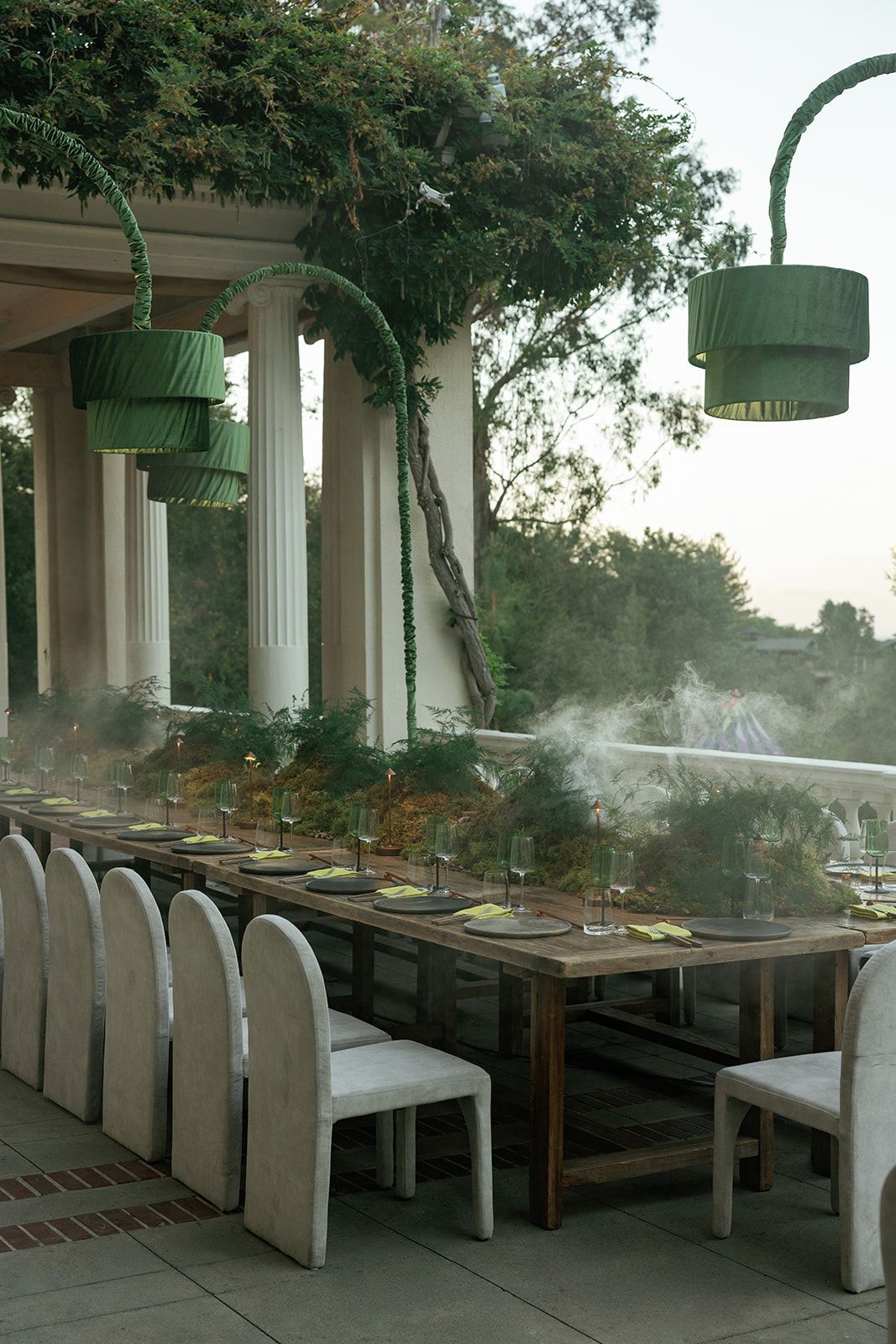 Elegant outdoor dining table set with glassware under green pendant lamps, surrounded by lush greenery and white columns.