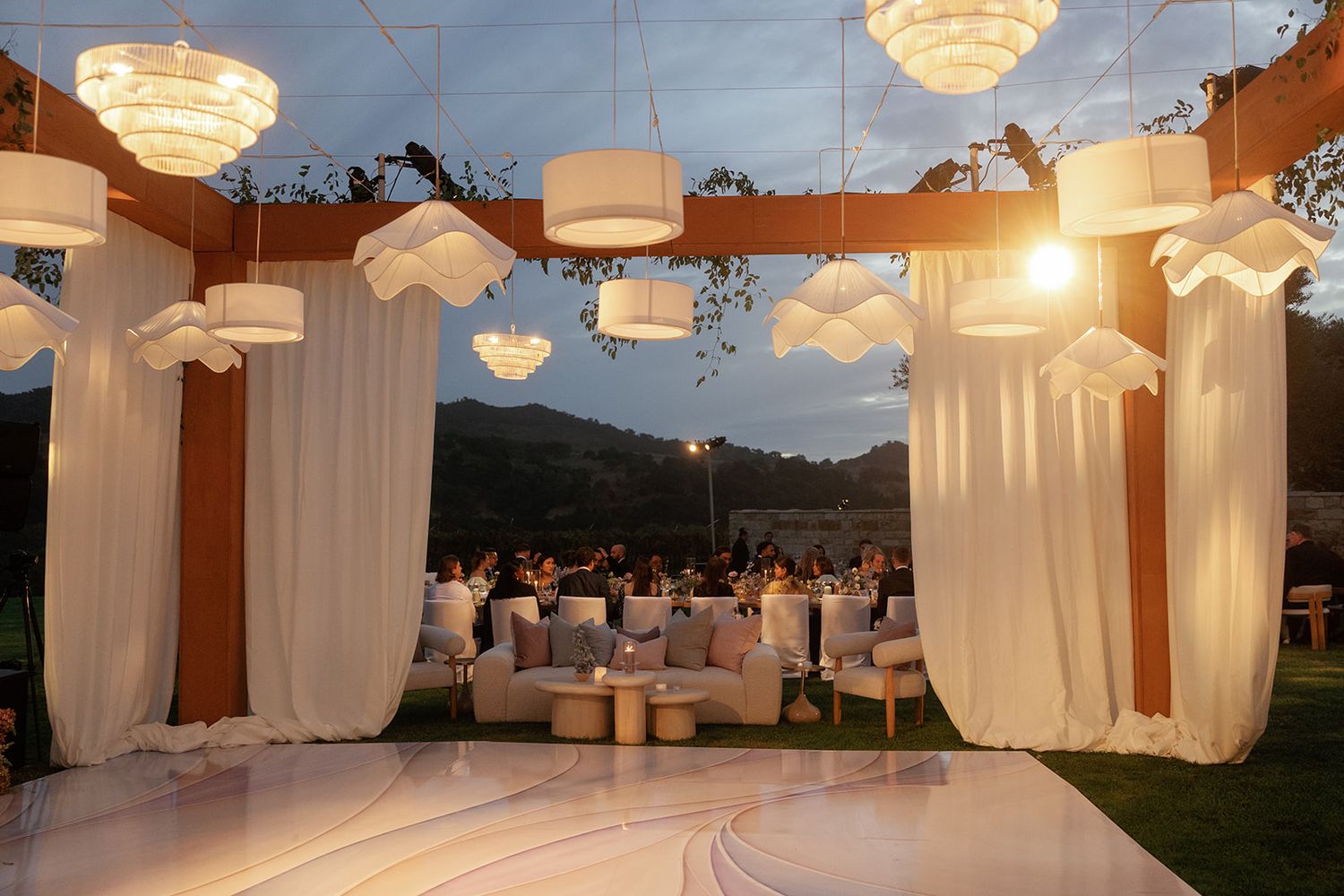 Outdoor wedding reception venue at dusk with white draping, chandeliers, and glowing pendant lights.