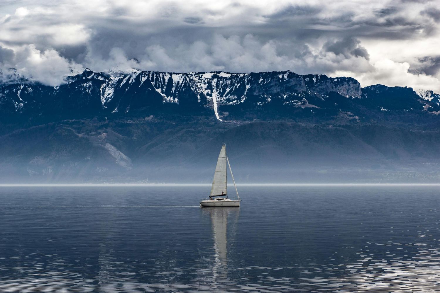 Lone sailboat on calm waters with dramatic snow-capped mountains rising in the background.