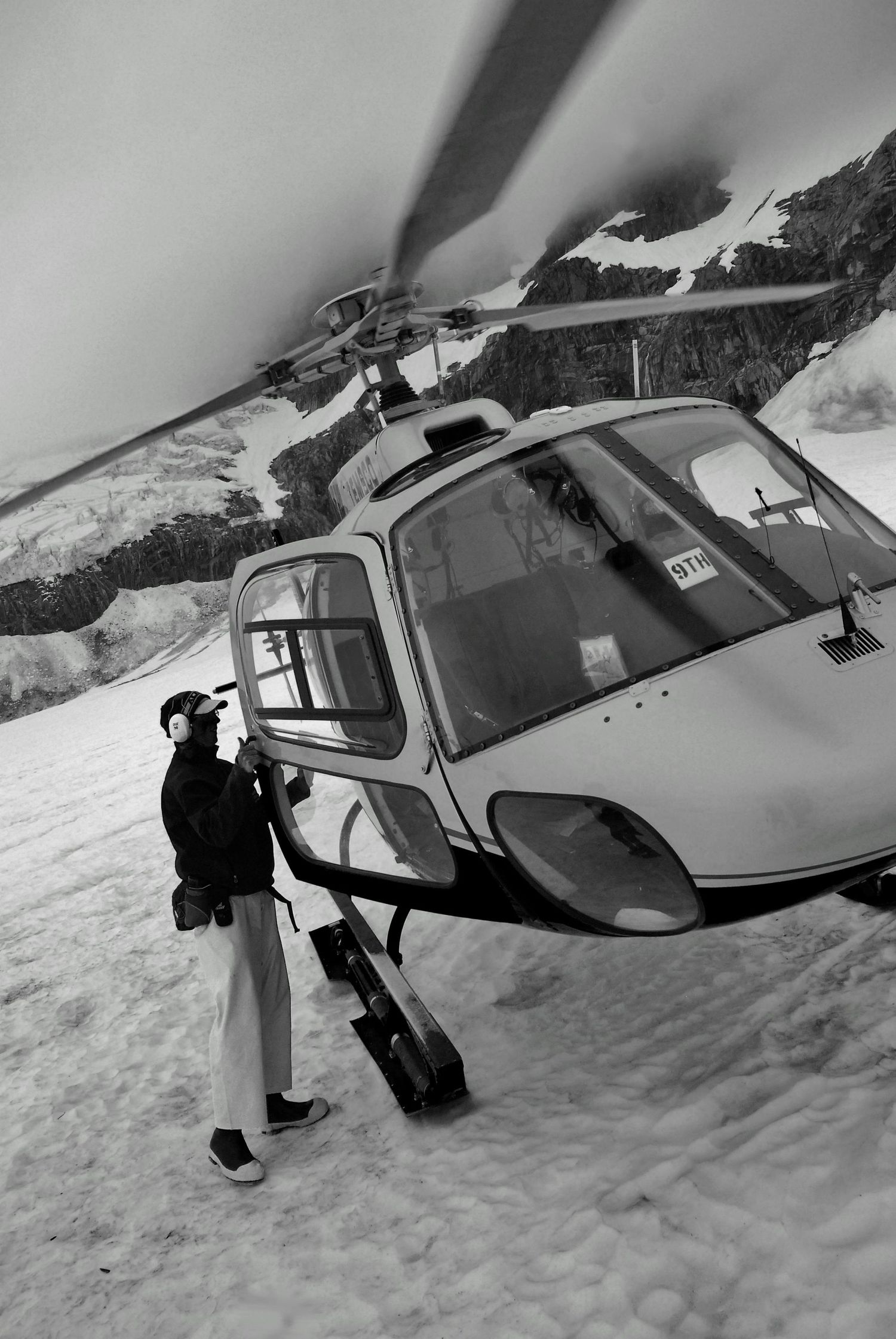A skier with camera equipment stands next to a helicopter parked on a snowy mountain slope in black and white.