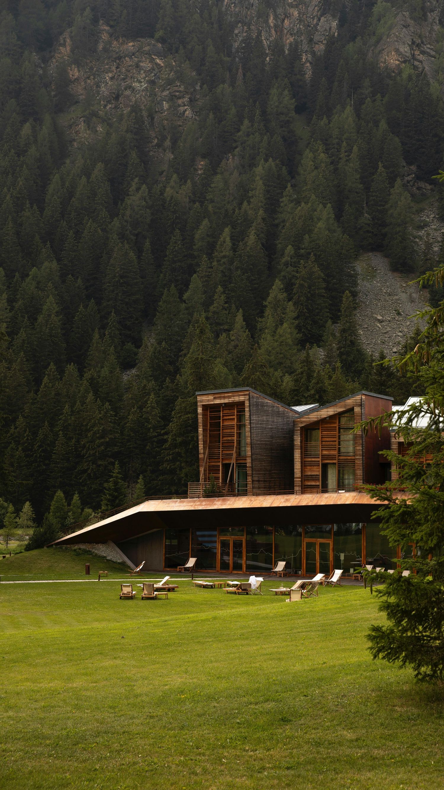 Modern wooden cabins nestled against a forested mountainside with Adirondack chairs on a lush green lawn.