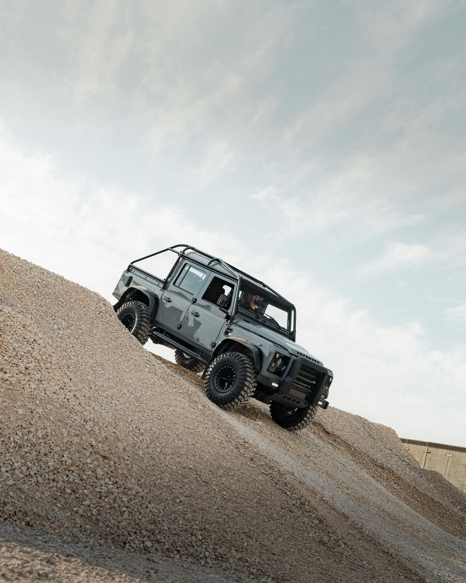 Land Rover Defender climbs a steep sandy slope against a cloudy sky.