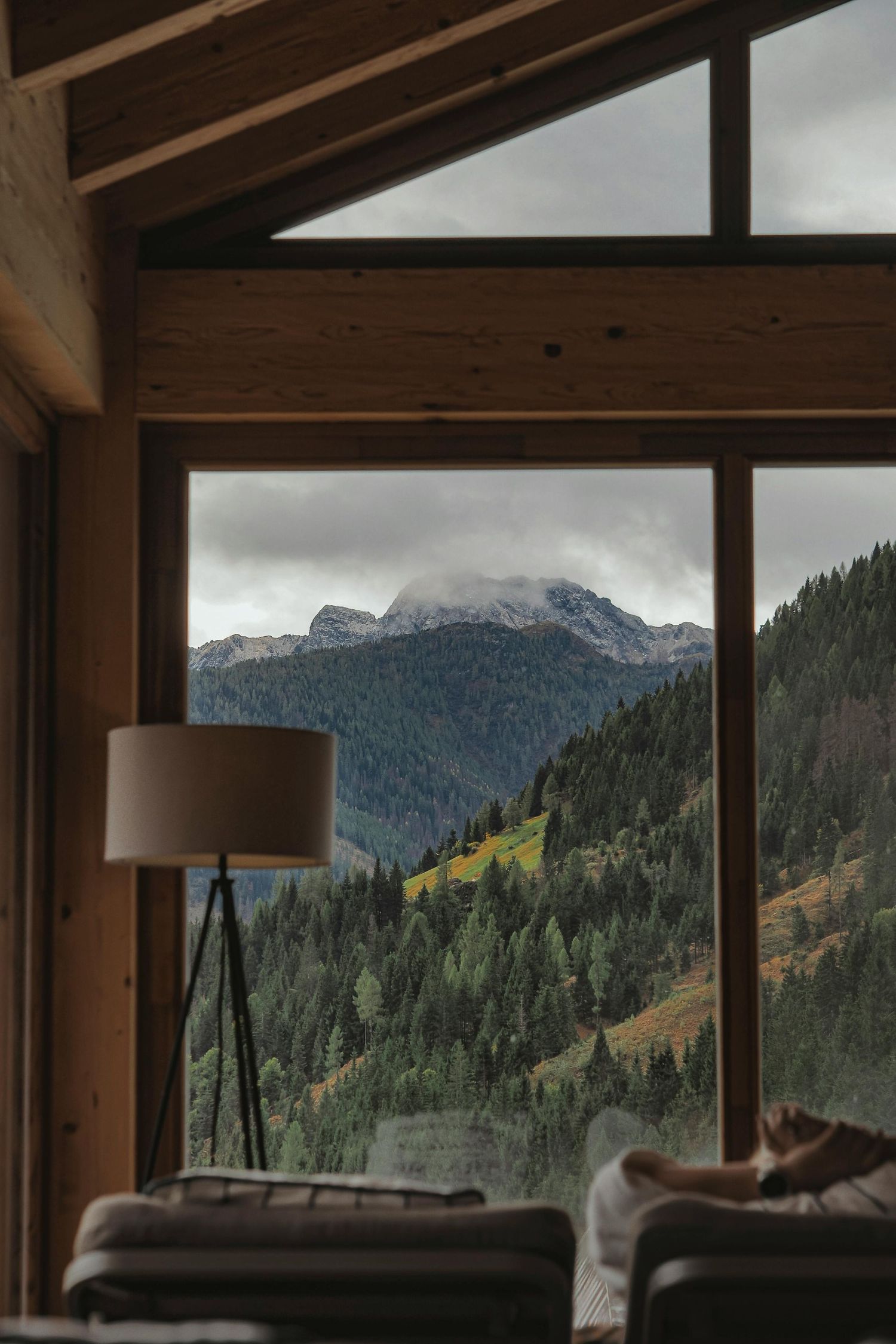 Panoramic mountain view through large windows of a modern cabin interior with exposed wooden beams and cozy seating.