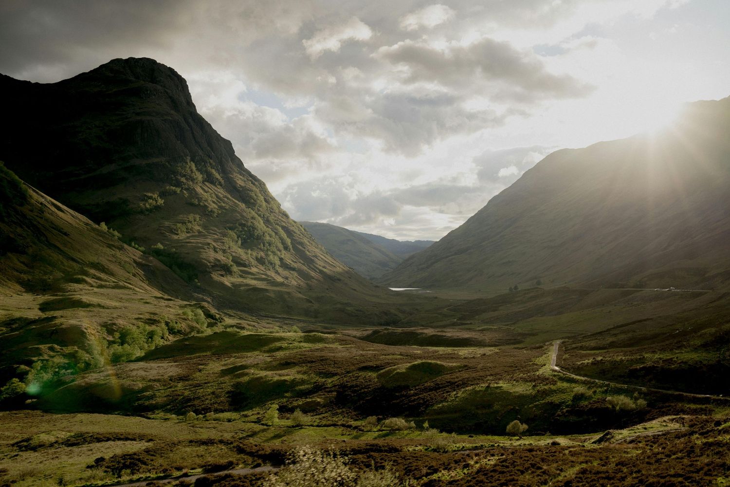 A dramatic valley landscape in Glen Coe, Scotland with sunlight streaming through cloudy skies.