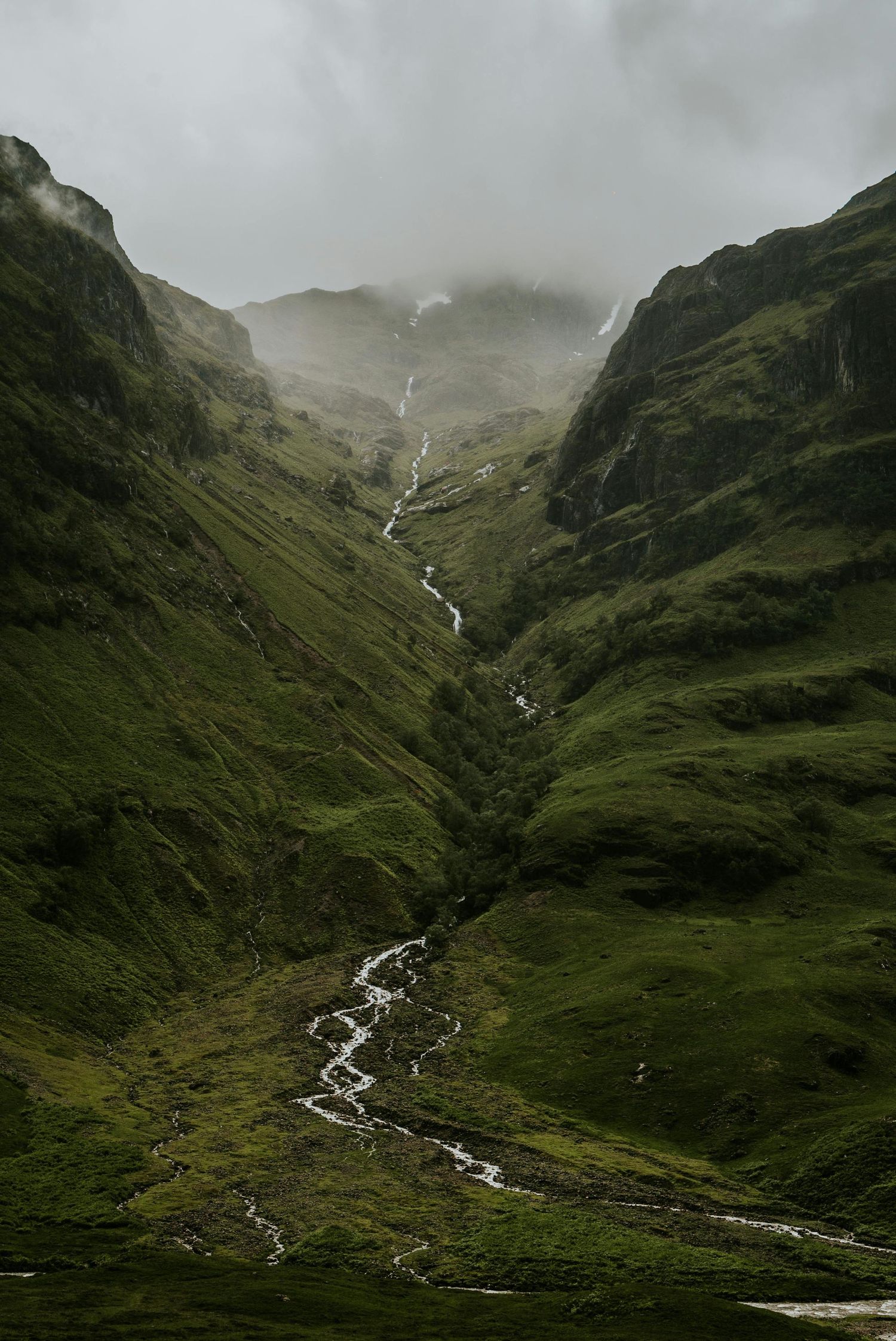 A cascading waterfall flows through a lush green valley between steep mountain slopes in misty conditions.